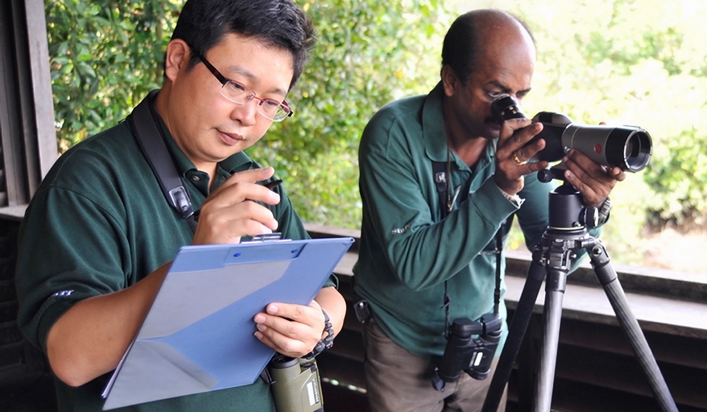 Two men in green shirts, one writing on a clipboard and one looking through a spotting scope on a tripod.